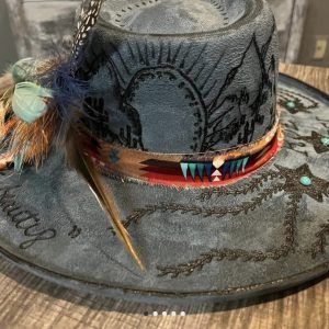 A handcrafted, wide-brimmed hat made of distressed blue-gray material. The hat features intricate black embroidery depicting desert scenes with cacti and mountains. A decorative hatband wraps around the base, showcasing a tan fabric with red, turquoise, and geometric patterns. Feathers in natural tones, with hints of teal and blue, are attached to the side of the hatband for added embellishment. Small turquoise accents are also placed on the brim, enhancing the hat's southwestern aesthetic.
